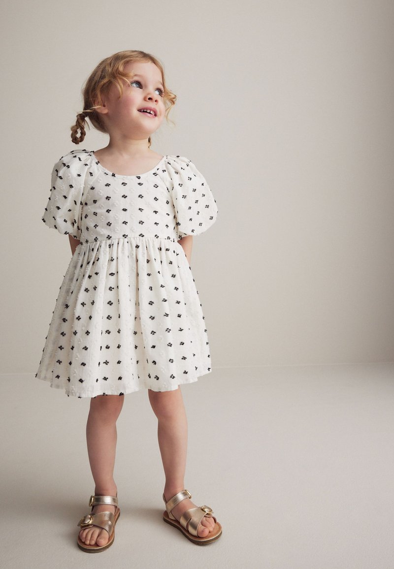 Robe blanche à manches bouffantes avec un petit motif floral noir ; coupe en A ; tissu texturé ; associée à des sandales dorées avec une boucle décorative.