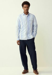 Šviesiai mėlynas ir baltas dryžuotas marškinėliai su ilgomis rankovėmis ir krūtine kišene, derinami su tamsiai mėlynomis kelnėmis ir rudais batais.