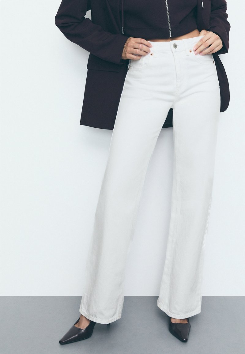 Personne portant un jean blanc taille haute à jambes larges, des talons noirs pointus, une veste noire zippée et un blazer sombre sur un fond uni.