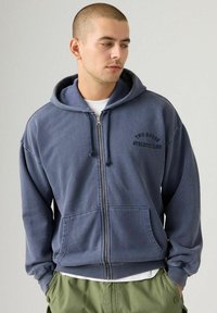 Joven con cabello corto vistiendo una sudadera azul con cremallera que dice "Two Horse Athletic Club", camiseta blanca y pantalones verdes, con las manos en los bolsillos.