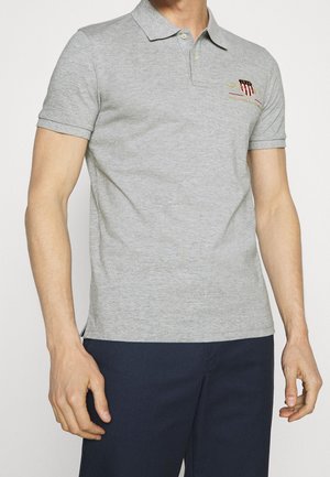 Homme portant un polo à manches courtes gris clair avec col et petit logo brodé sur la poitrine, associé à un pantalon bleu foncé.