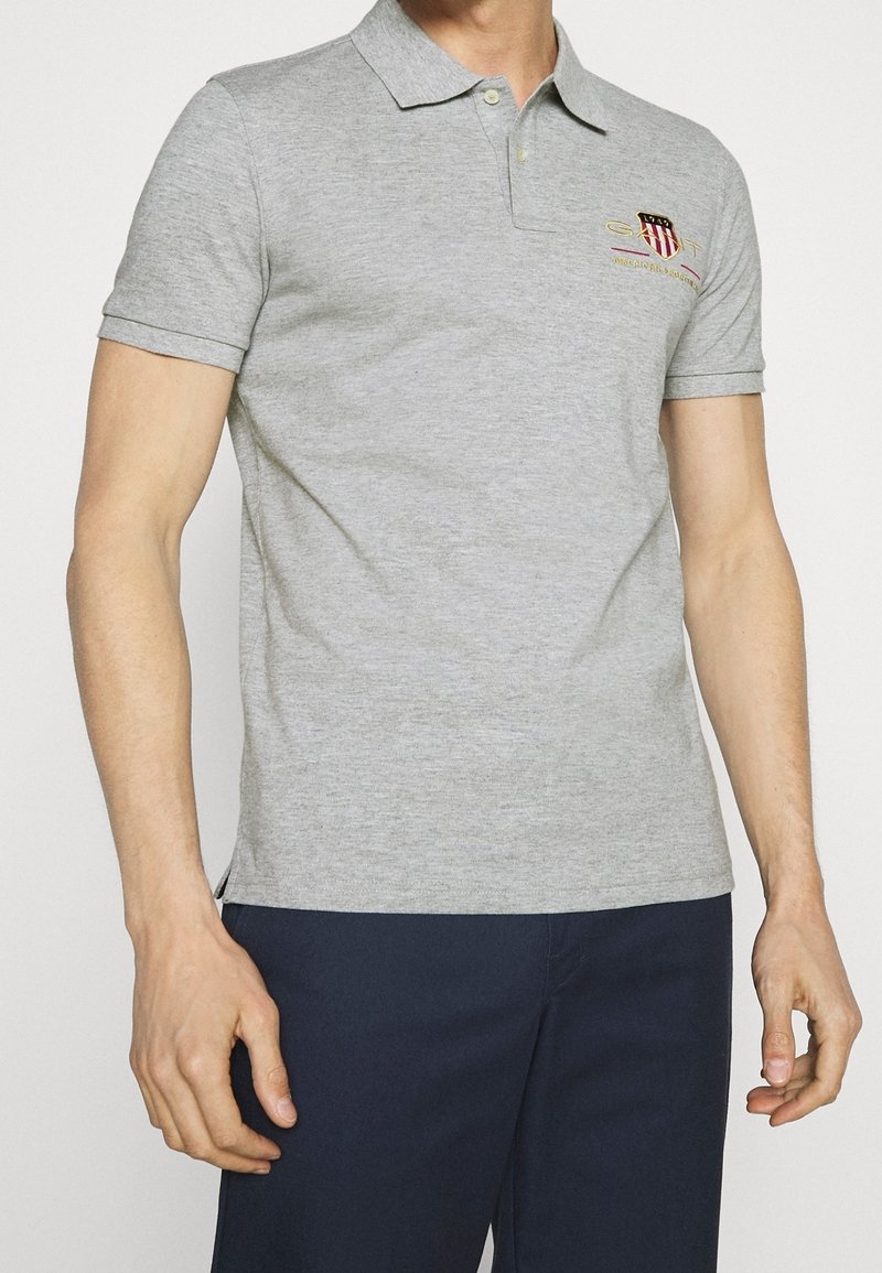 Homme portant un polo à manches courtes gris clair avec col et petit logo brodé sur la poitrine, associé à un pantalon bleu foncé.