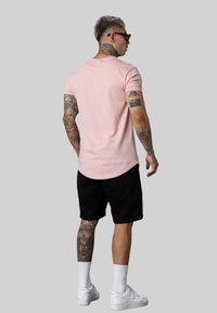 Helles rosa, tailliertes T-Shirt mit kurzen Ärmeln, kombiniert mit schwarzen Shorts. Weiße Sneaker und Kniesocken. Sichtbare Tattoos an Armen und Beinen.