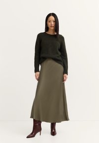 Dunkelgrüner, gestrickter Pullover, kombiniert mit einem olivgrünen, satinartigen Maxikleid. Braune, spitz zulaufende Stiefeletten komplettieren das Outfit.