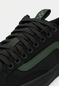 Crne tenisice s gornjim dijelom od teksturirane tkanine i zelenim akcentom, okruglim crnim rupama za vezanje i odgovarajućim crnim vezicama.