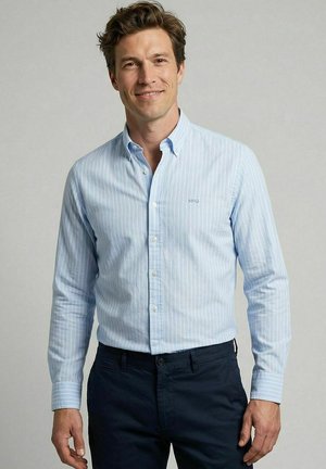 Man draagt een lichtblauw gestreept overhemd met knopen en een donkere broek, staat tegen een effen lichtgrijze achtergrond en glimlacht licht.