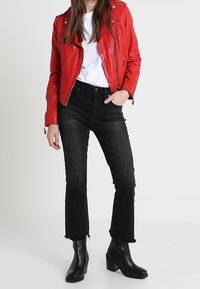 Vrouw in een rode leren jack, witte T-shirt, zwarte hoge taille jeans met franjes en zwarte haklaarzen, staand met een hand in haar zak.