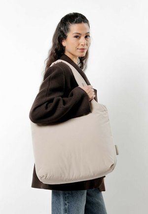 Sac fourre-tout en tissu beige avec une grande forme douce. Bande réglable, texture lisse et design minimaliste. Porté sur un manteau sombre.