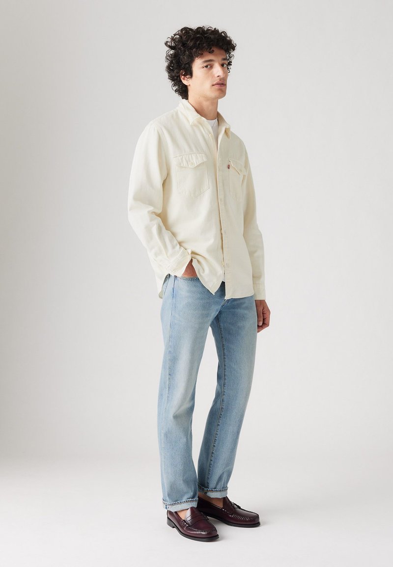 Hombre de pie con las manos en los bolsillos, vistiendo una camisa de botón color crema, jeans azul claro y mocasines burdeos oscuros sobre un fondo blanco.