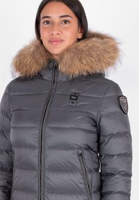 Grijze puffy jas met een afneembare bruine bontkap. Heeft een ritssluiting aan de voorkant, zijzakken en een logo-opdruk op de linker mouw.