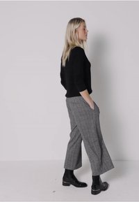 Pull en laine noir associé à un pantalon large à carreaux gris. Le mannequin porte des bottines noires avec une touche dorée. Design simple et moderne.