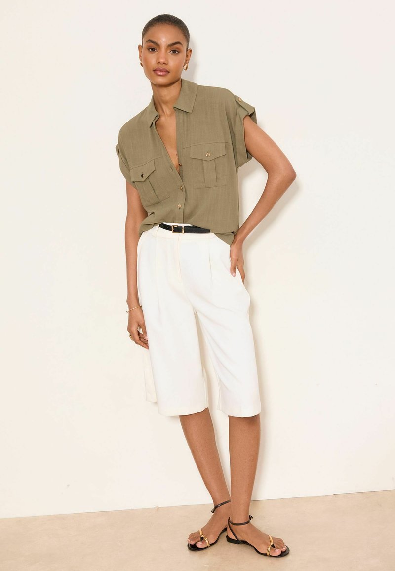 Frau trägt ein olivfarbenes, kurzärmliges Hemd mit Brusttaschen, weiße, hoch taillierte, weit geschnittene Shorts, einen schwarzen Gürtel und schwarze Sandalen.