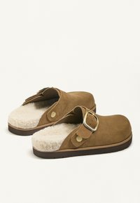 Clogs din suede maro cu o talpă interioară moale din blană sintetică. Prezintă o curea lată, o cataramă din alamă și o talpă din cauciuc cu un underside texturat pentru aderență.