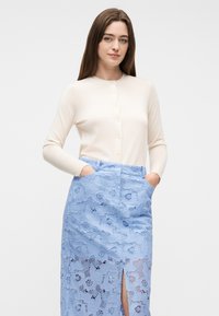 Γυναίκα με μακριά καστανά μαλλιά, που φοράει κρεμ ζακέτα με κουμπιά και φούστα από μπλε δαντέλα με ψηλή μέση και τσέπες, στέκεται μπροστά σε απλή φόντο.