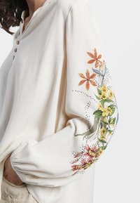 Krämfärgad blus med långa ärmar och träknappar. Har detaljerad blommönsterbroderi i orange, gult och grönt på ärmarna.