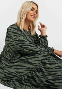 Grön mönstrad klänning i en texturerad väv, med ett lager-på-lager design, långa ärmar och elastiska manschetter med ett svart abstrakt mönster.