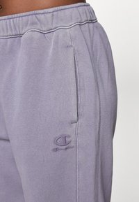 Gaiši violeto trenniņas, kas izgatavotas no mīksta kokvilnas maisījuma. Iezīmes ietver elastīgu jostasvietu, sānu kabatas un logo izšuvumu uz augšstilba.