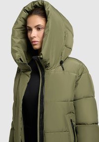 Olīvu zaļš pildīts mājoklis ar lielu kapuci, frontālo rāvējslēdzēju un sānu kabatām. Gluda tekstūra, pildījums no pūkām un kontrastējošs melns rāvējslēdzējs.