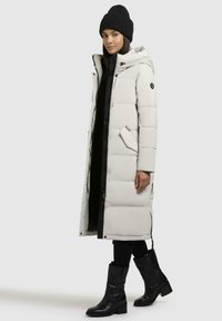 Lang, puffy kåpe i lys grå med hette, svart glidelås og lommer. Bært over et svart antrekk og kombinert med svarte, klumpete hæl-støvler.