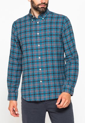 Uomo che indossa una camicia a quadri blu e grigia a maniche lunghe con taschino sul petto, abbinata a pantaloni scuri su sfondo bianco.