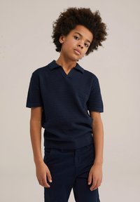 Navy modrá polokošeľa s krátkymi rukávmi, textúrovaným vzorom a výstrihom do V, v kombinácii s tmavými nohavicami. Hladký povrch látky.