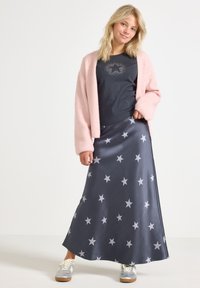 Jeune femme portant un cardigan rose, une jupe bleu marine à motifs étoiles, une chemise noire à motifs étoiles, et des baskets claires, se tenant devant un fond blanc.