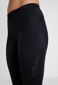 Leggings atléticos pretos feitos de um tecido elástico e que absorve a umidade. Apresentam costuras cosidas e um logotipo impresso a preto de lado.