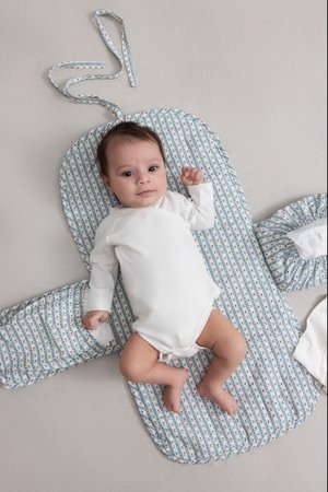 Un bébé dans une combinaison blanche repose sur un matelas à langer rembourré avec un motif floral bleu et blanc et des soutiens latéraux, sur une surface grise lisse.