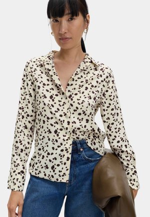 Vrouw met een crèmekleurig overhemd met zwarte abstracte vlekken, blauwe jeans, gouden oorringen, en een bruine leren jas over haar arm houdend.