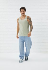 Light green cotton tank top with a scoop neck, featuring a small black logo on the chest, paired with loose blue jeans and sneakers.