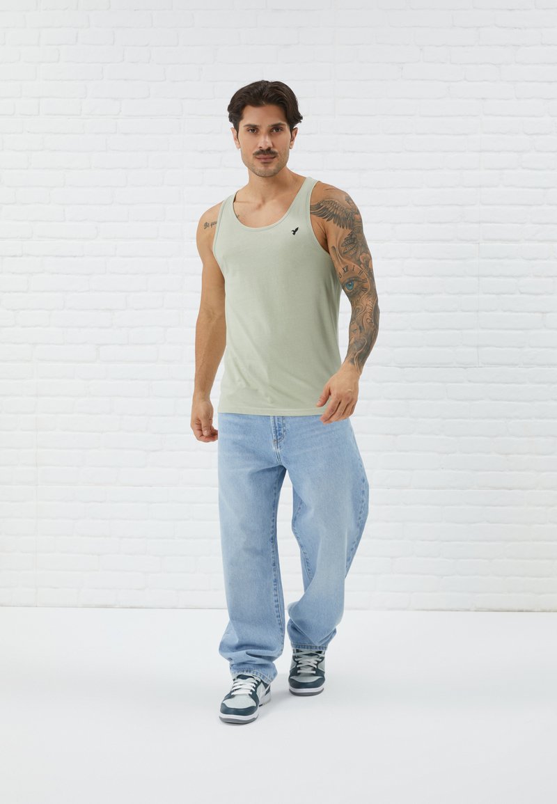 Light green cotton tank top with a scoop neck, featuring a small black logo on the chest, paired with loose blue jeans and sneakers.