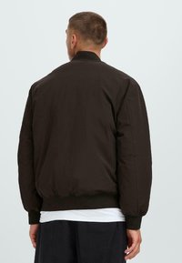 Dark brown bomber jacket with a ribbed collar and cuffs. Features a smooth, slightly padded material and a relaxed fit design.