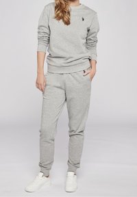 Person trägt einen hellgrauen Sweatshirt und passende Sweatpants mit weißen Sneakers und steht vor einem schlichten hellen Hintergrund.