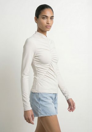 Donna con capelli raccolti indietro indossa una maglietta beige arricciata a maniche lunghe e pantaloncini di jeans azzurri, in piedi su uno sfondo chiaro e uniforme.