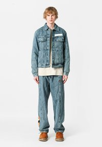 Chaqueta de mezclilla con un lavado azul claro, bolsillos en el pecho y una etiqueta blanca. Combinada con pantalones de mezclilla azul claro que presentan un detalle de parche.