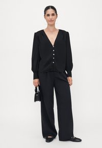 Blusa nera con dettagli di pence e bottoni, caratterizzata da maniche lunghe a sbuffo. Abbinata a pantaloni neri a gamba larga e scarpe nere flat.