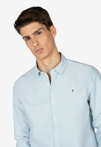 Camisa de botones de color azul claro con finas rayas blancas, diseño con cuello, que presenta un pequeño logo en color navy en la zona izquierda del pecho.