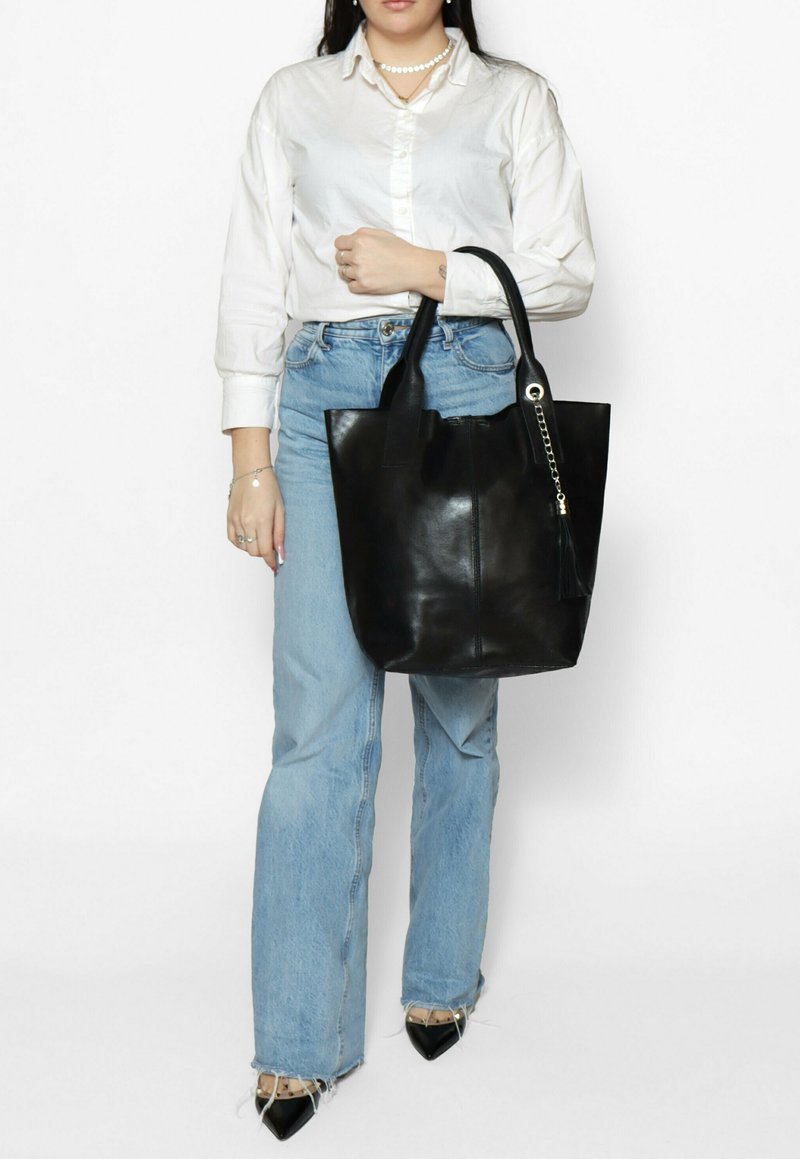 Sac cabas en cuir noir à texture lisse, avec deux anses robustes, une fermeture éclair et un accent de pompon. Mis en scène sur un fond uni.