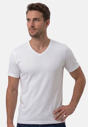 Homme aux cheveux bruns courts et à la barbe, portant un t-shirt blanc uni à col en V, debout, la main sur la hanche, regardant sur le côté.