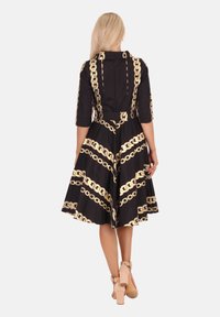 Frau mit langen blonden Haaren, die ein schwarzes Kleid mit goldenen Kettenmustern und beigen High-Heels mit Knöchelriemen trägt, steht mit dem Rücken zur Kamera.