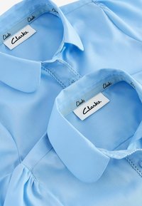 Blusa azzurro chiaro con colletto classico, dettagli cuciti e bottoni, con etichette del marchio visibili all'interno del colletto.