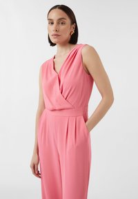 Roze mouwloze jumpsuit met een gedrapeerde halslijn, een ceintuurlijn in de taille en zijzakken; gemaakt van een gladde stof met een subtiele glans.