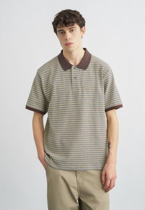 Joven con cabello corto y castaño, vistiendo una camiseta polo de rayas con cuello marrón y pantalones beige, de pie con las manos en los bolsillos.