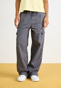 Pantalon cargo gris à coupe décontractée, doté de poches latérales et d'une taille à cordon. Associé à des baskets blanches sur un sol jaune.