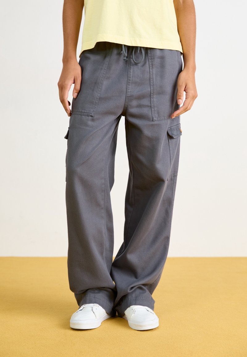 Pantalon cargo gris à coupe décontractée, doté de poches latérales et d'une taille à cordon. Associé à des baskets blanches sur un sol jaune.