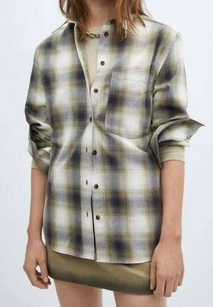 Woman wearing rolled-up sleeves plaid shirt in muted green, black, and white tones over a beige long-sleeve top and olive skirt.