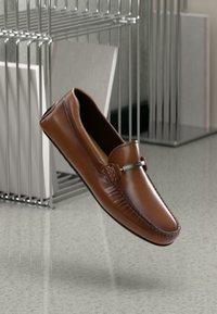 Loafers din piele maro cu cusături decorative, o textură netedă și un accent argintiu pe partea superioară. Ușori și fără șnururi.