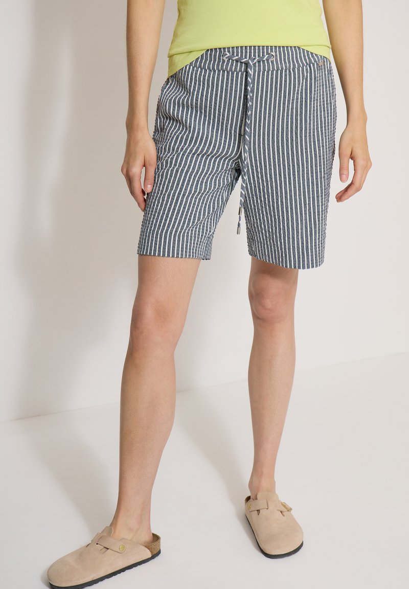 Personne portant un short rayé vertical bleu et blanc avec un cordon, un t-shirt jaune et des chaussures beiges sans lacets.