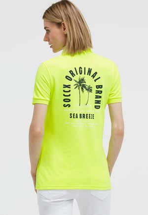 Frau mit kurzen blonden Haaren, die ein neongelbes Polohemd mit schwarzem Palmenmotiv und dem Schriftzug „SOCCX ORIGINAL BRAND SEA BREEZE“ auf dem Rücken trägt.