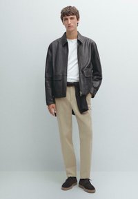 Veste en cuir noir avec deux poches avant, portée sur une chemise blanche. Un pantalon beige et des chaussures en daim marron foncé complètent la tenue.